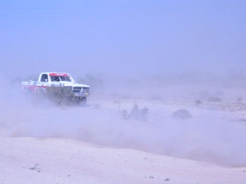 1990 Ford Bronco - white and red off-road race truck drives through a hazy veil of dust over sandy desert ground.