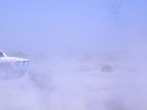 1990 Ford Bronco - white race truck with BFGoodrich decals emerges from a heavy desert dust cloud near a power tower.