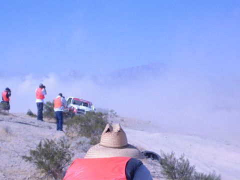 1990 Ford Bronco - spectator in a straw hat and red vest watches a race truck and orange-vested photographers amid desert dust.