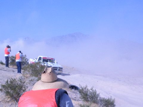 1990 Ford Bronco - straw-hatted spectator and orange-vested photographers watch a race truck approach through desert dust.