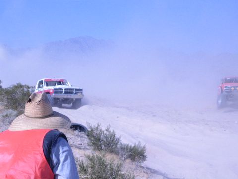 1990 Ford Bronco - white truck and a red truck approach a straw-hatted spectator across a dusty sandy desert course.