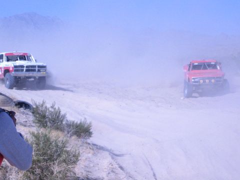 1990 Ford Bronco - photographer in the foreground films two off-road race trucks, one white and one red, churning through dust on a desert course.