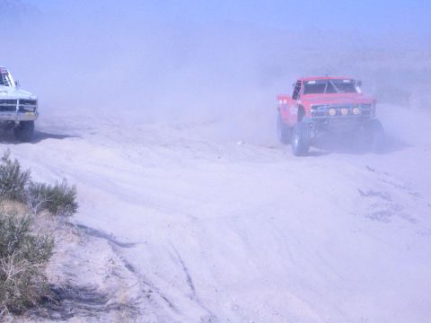 1990 Ford Bronco - red off-road race truck climbs a sandy berm trailing thick dust, with a white race truck visible at left.