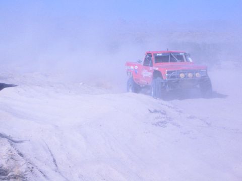 1990 Ford Bronco - red off-road race truck marked 800 crests a sandy rise enveloped in desert dust.