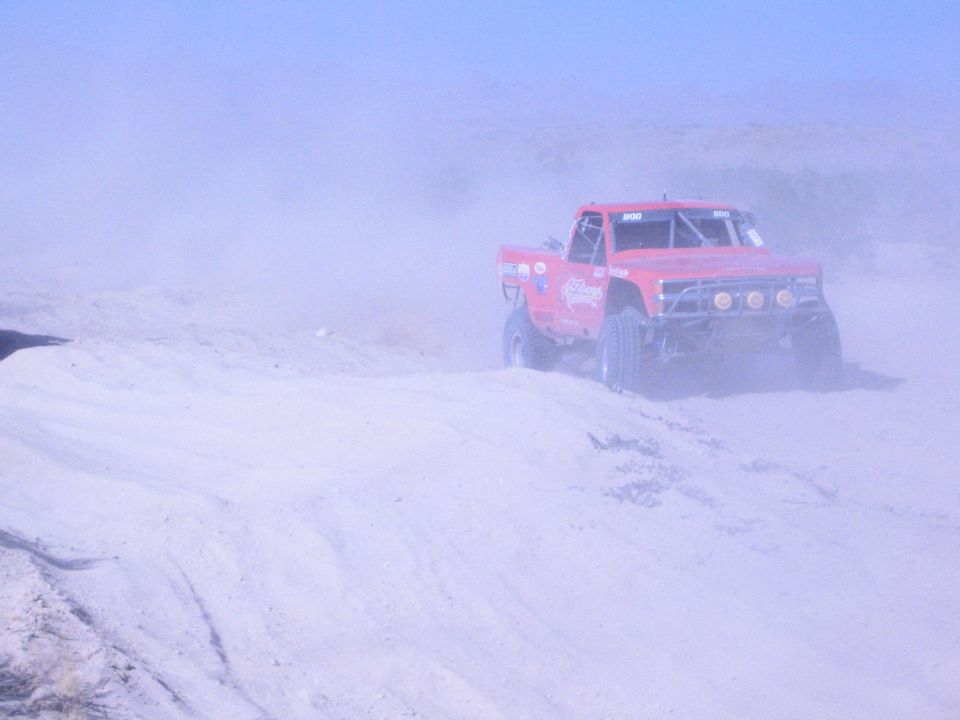 Red number 800 off-road race truck cresting a sand berm in heavy dust