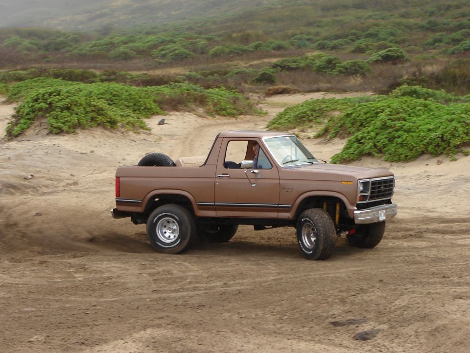 Brown 1985 Ford Bronco driving on sand, spare tire in the bed. Coastal hills in background
