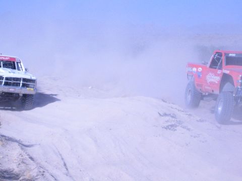 1990 Ford Bronco - white race truck and a red race truck travel a dusty desert track, the red truck lettered along its side.
