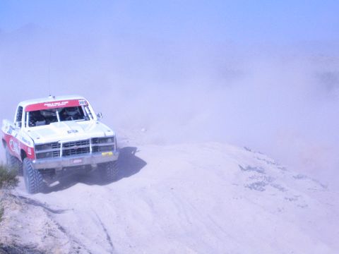 1990 Ford Bronco - white-and-red off-road race truck rounds a sandy berm, raising a cloud of dust against a clear sky.