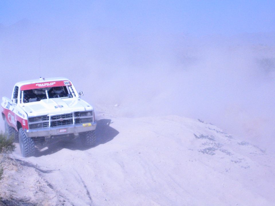 White and red off-road race truck rounding a sandy desert berm in dust