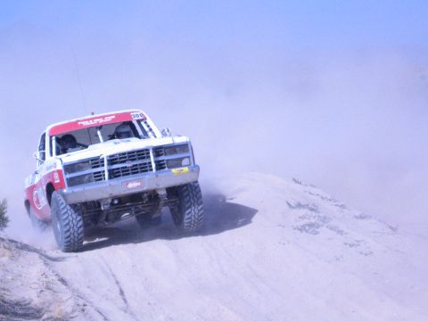 1990 Ford Bronco - white-and-red off-road race truck climbs a sand berm head-on, kicking up dust under a blue sky.