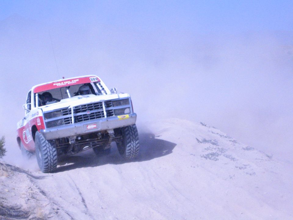 Front view of white and red off-road race truck climbing sand in desert dust