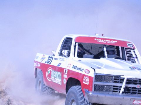 1990 Ford Bronco - Side view of off-road race truck 301 with a window safety net, lettered California Tow and BFGoodrich, driving through dust.