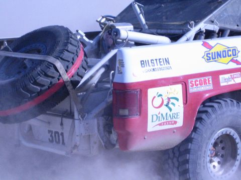 1990 Ford Bronco - Rear quarter of off-road race truck 301 with a chained spare tire and Sunoco, DiMare and Bilstein decals, kicking up dust.