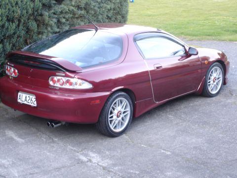 1993 Mazda MX-3 - The rear of a maroon 1993 Mazda MX-3 coupe is shown, a prominent spoiler and dual exhaust pipes.