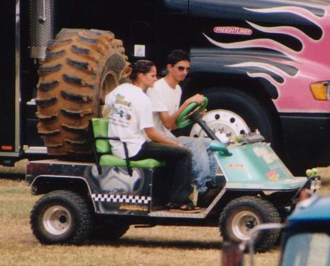 1987 Ford Bronco - A light green golf cart carries two passengers, with a large tire and a Freightliner truck visible behind them.