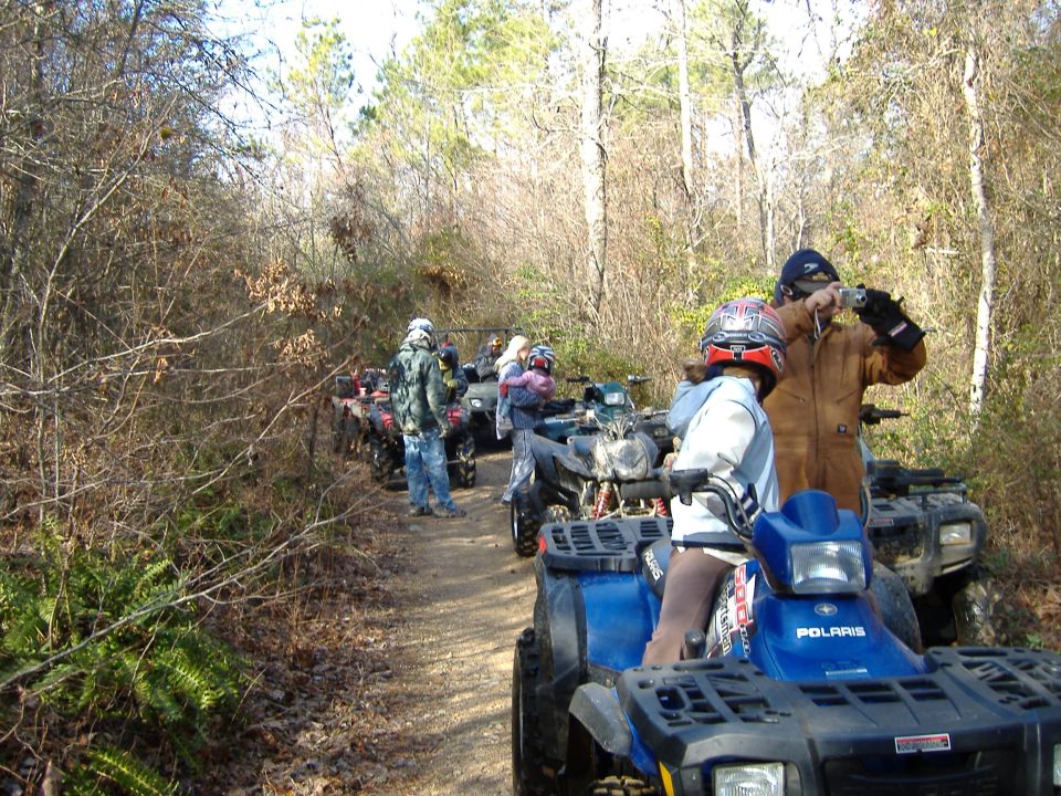 Group of people on ATVs lined up on a dirt path in a wooded area. One person is taking a photo