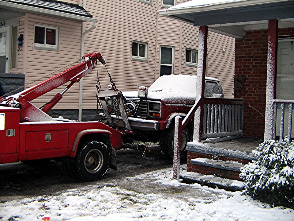 Red tow truck hooking up a snow-covered red pickup in a driveway in winter