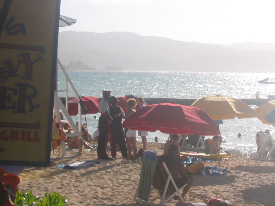Beach scene with people under umbrellas, ocean in background. Sign on left says 'HEAVY BURGER GRILL'
