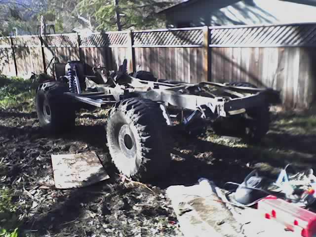 Stripped truck frame with axles and large tires in a backyard by a wood fence