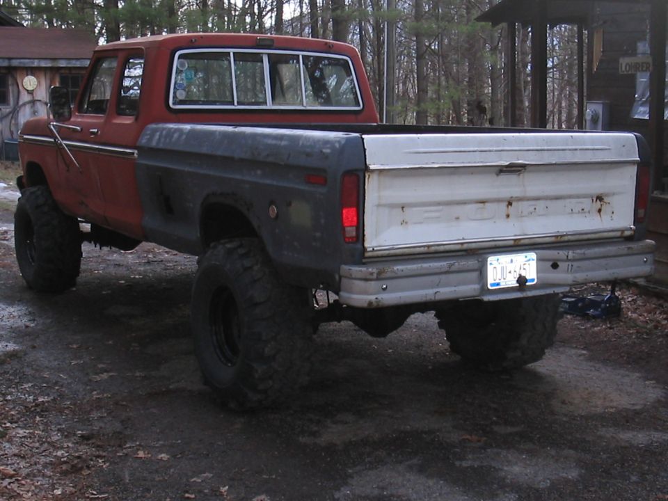 Lifted square-body Ford 4x4 pickup, red cab and gray bed, rear three-quarter on gravel