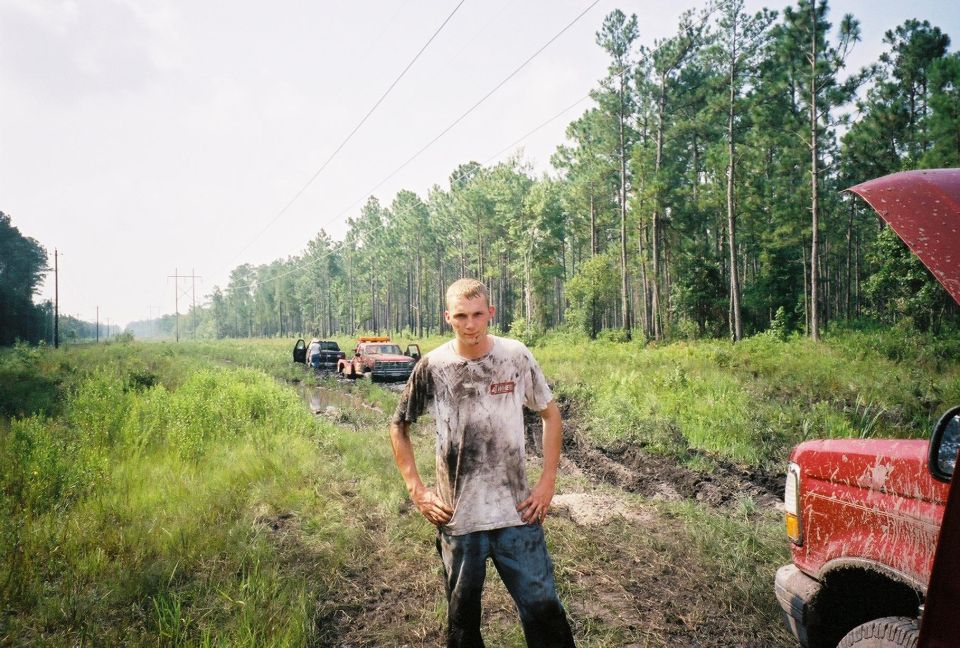 Muddy man on off-road trail; red vehicle with hood up at right, tow truck stuck in mud in background