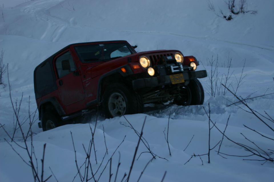 Red 2003 Jeep TJ driving through deep snow, headlights on