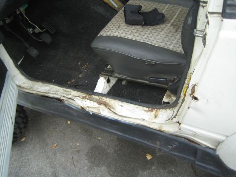1987 Suzuki Samurai - The interior of a white vehicle shows significant rust and damage along the driver's side door sill.