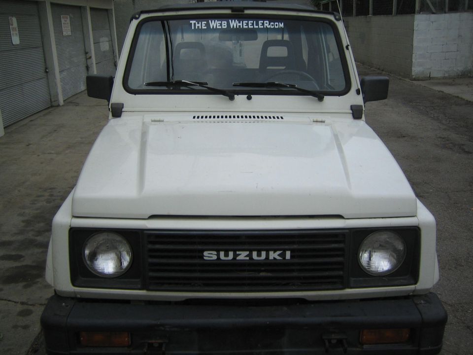 White Suzuki Samurai front view, grille reads SUZUKI. Sticker on windshield says THE WEB WHEELER.COM