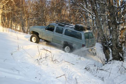 2003 Jeep TJ - A light blue SUV navigates a snowy trail, kicking up snow as it drives.