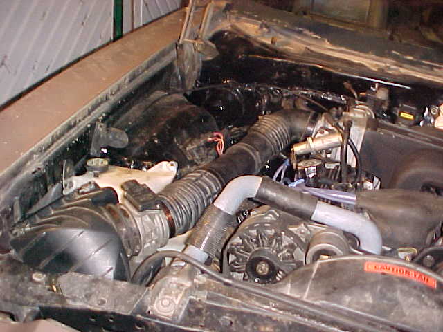 Engine bay with black corrugated intake hose, alternator, and orange CAUTION FAN label