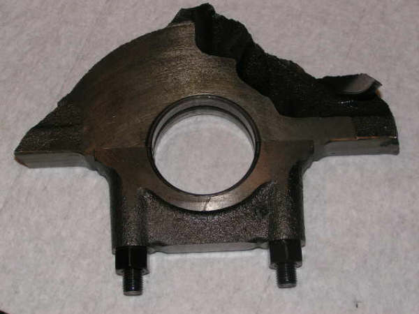 Damaged metal engine part with a circular opening and two bolts, resting on a textured surface