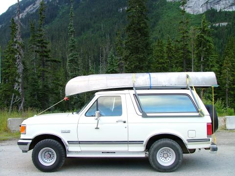 1988 Ford Bronco - A white Ford Bronco is parked on a dirt road, carrying a canoe on its roof rack, with a mountainous forest in the background.
