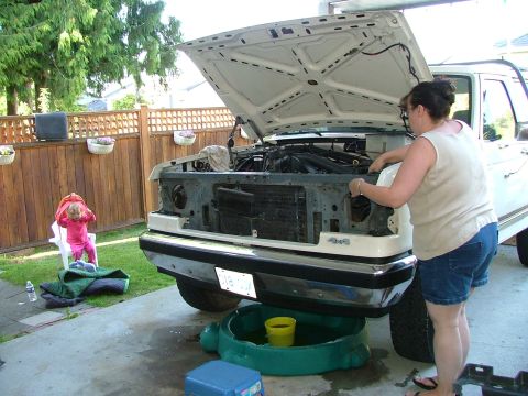 1988 Ford Bronco - A woman works on the engine of a white Ford truck in a driveway, with a child watching from a chair.