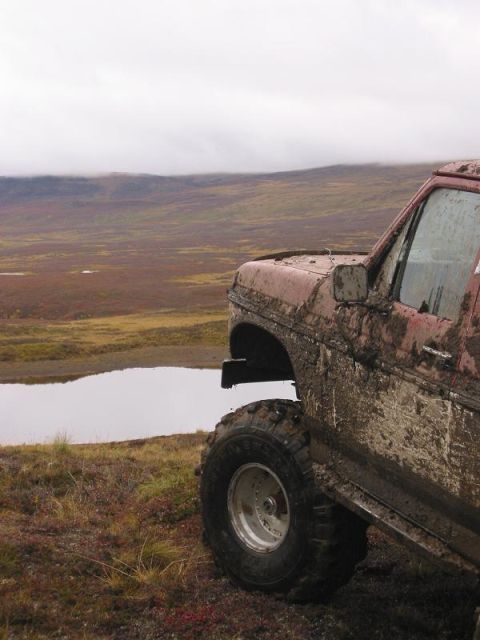 1985 Ford Bronco - A heavily mud-covered red SUV with large off-road tires sits on a hill overlooking a vast, barren landscape.
