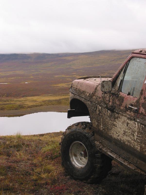 Muddy maroon lifted Bronco front corner on tundra by a pond under gray skies