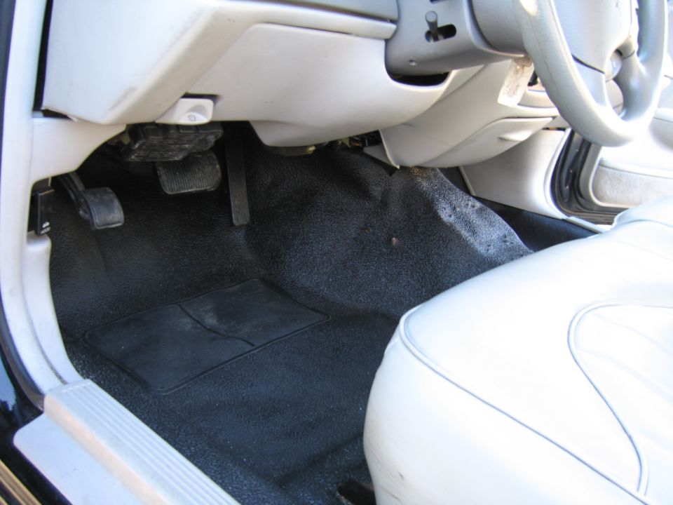 Car's driver side footwell with black carpet, floor mat, and pedals visible. White seat is on the right