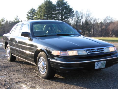 1991 Ford LTD Crown Victoria - The front end of a black 1991 Ford Crown Victoria is visible, parked on a gravel surface with trees in the background.
