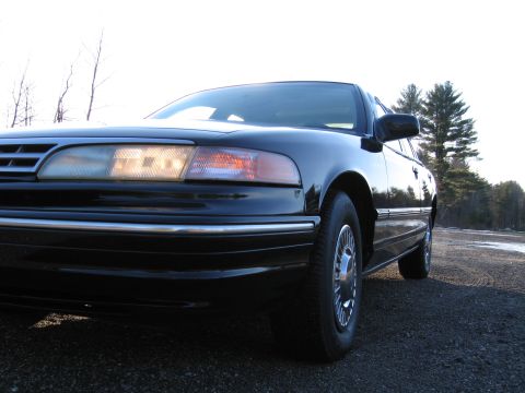 1991 Ford LTD Crown Victoria - A black sedan is viewed from the front quarter angle, parked outdoors on a gravel surface.