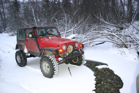 2003 Jeep TJ - A red Jeep TJ navigates a snowy path next to a partially frozen stream, with snow-covered trees in the background.