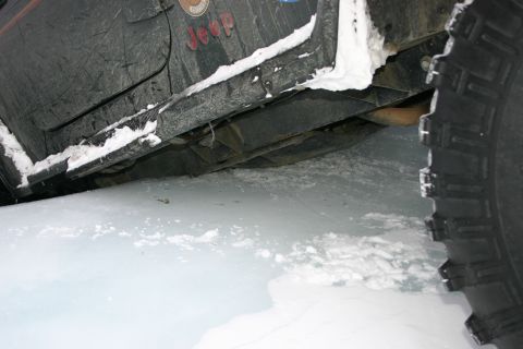 2003 Jeep TJ - The side and undercarriage of a Jeep TJ are visible, partially covered in snow, as it sits on a frozen surface.