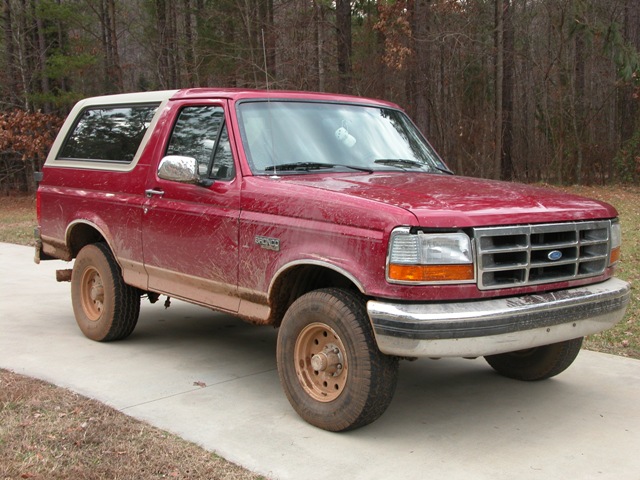 Mud-splattered red Ford Bronco SUV with tan top, 3/4 front view on a driveway among winter trees