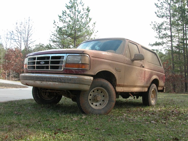 Mud-covered dark red Ford Bronco SUV parked on grass, low front three-quarter view with pine trees behind