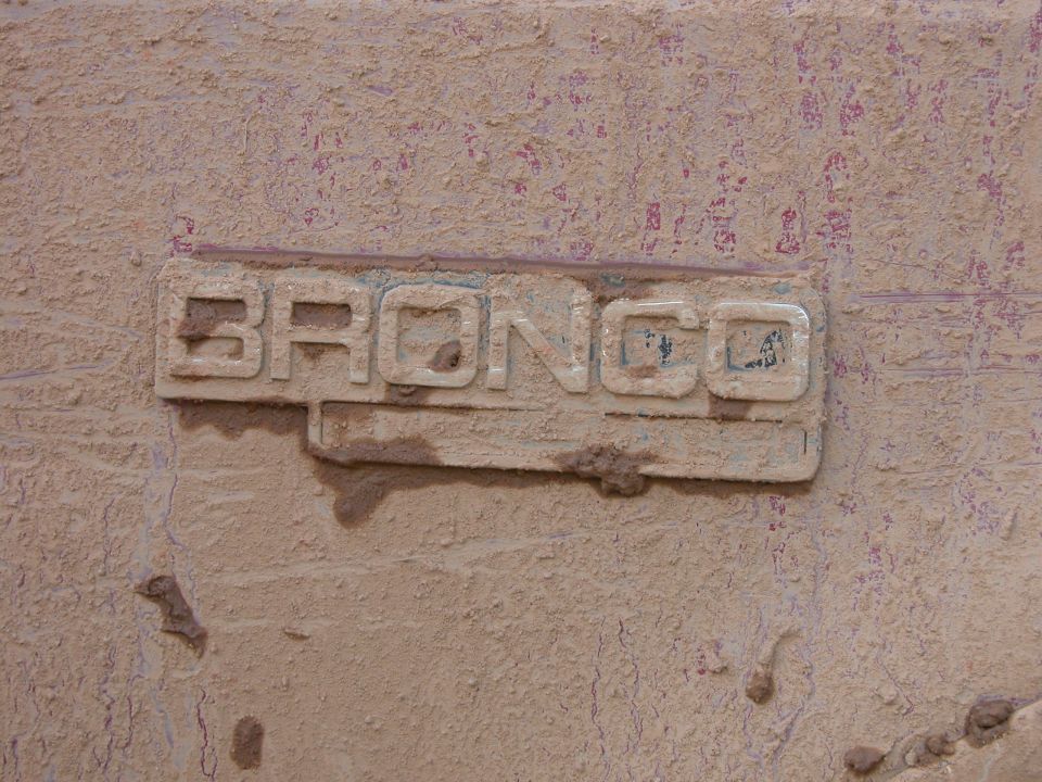 Weathered chrome 'BRONCO' fender emblem on a tan paint-flaked body panel