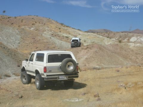 1988 Ford Bronco - Two white SUVs navigate a dusty, arid landscape under a clear blue sky.