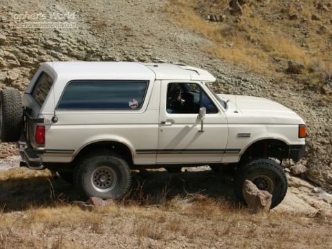 1988 Ford Bronco - A white Ford Bronco navigates a rocky, off-road path with a spare tire mounted on the rear.