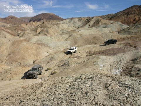1988 Ford Bronco - Multiple SUVs navigate a dusty trail through a barren, hilly desert landscape under a clear blue sky.