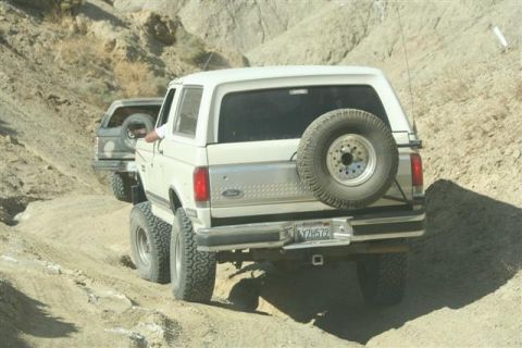 1988 Ford Bronco - Two Ford Broncos navigate a dusty off-road trail in a desert landscape.