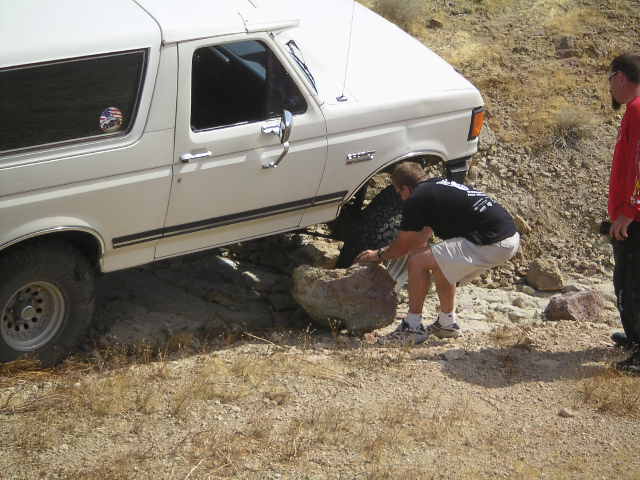 White 1988 Ford Bronco with its front tire stuck on a rock, a man is trying to move the rock