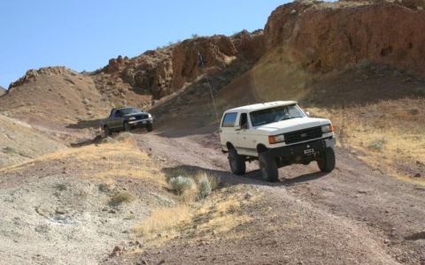 1988 Ford Bronco - Two off-road vehicles, a white Ford Bronco and a black pickup truck, navigate a dusty desert trail under a clear blue sky.