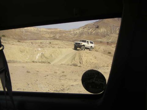 1988 Ford Bronco - A white SUV navigates a dusty, arid terrain with mountainous backdrops, viewed from the interior of another vehicle.